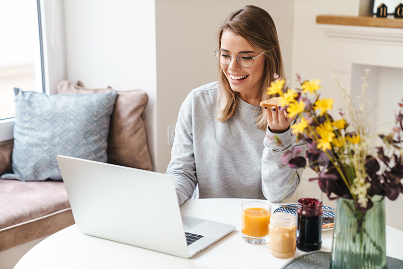 cheerful-woman-working-from-home A woman eating breakfast and looking at her laptop.