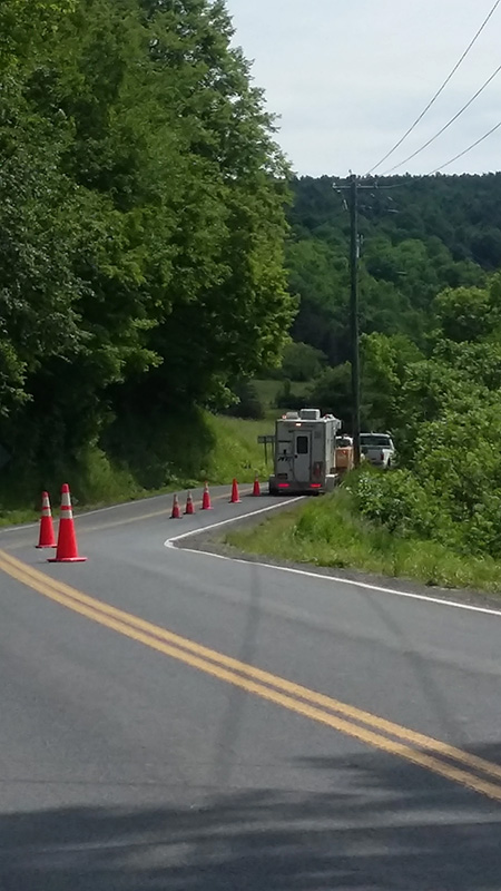 A utility truck and crew vehicle stop traffic on a curving country road using orange cones, with wooded hills and power lines in the background.